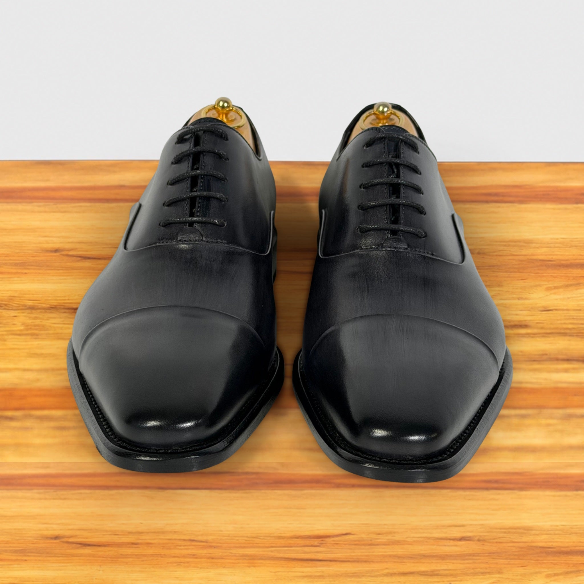 A pair of men's Gray leather oxford dress shoes with a cap toe, shown from the front on a wooden surface against a light gray background.