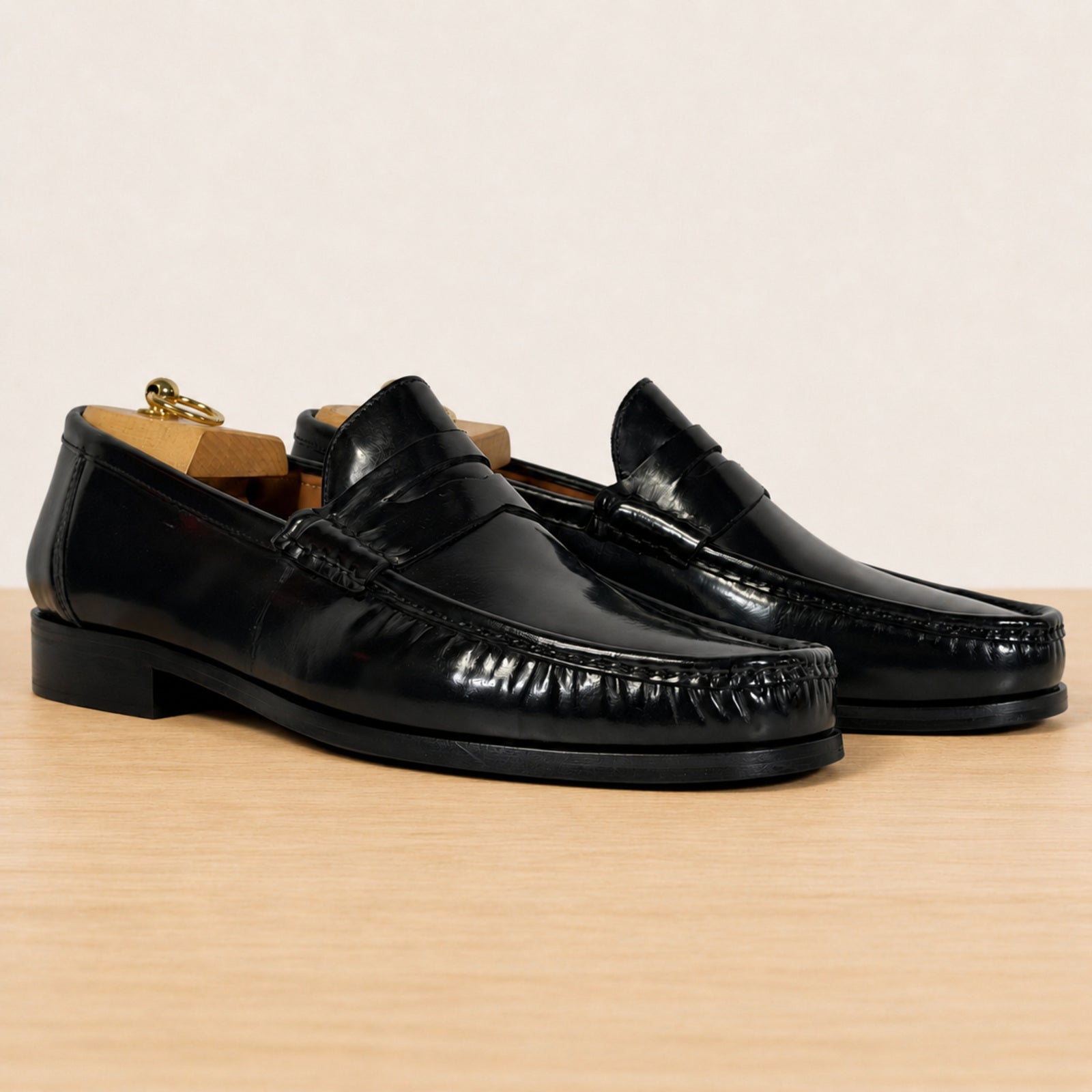 A pair of black leather penny loafers with a glossy finish, displayed on a light wood surface against a warm beige background.