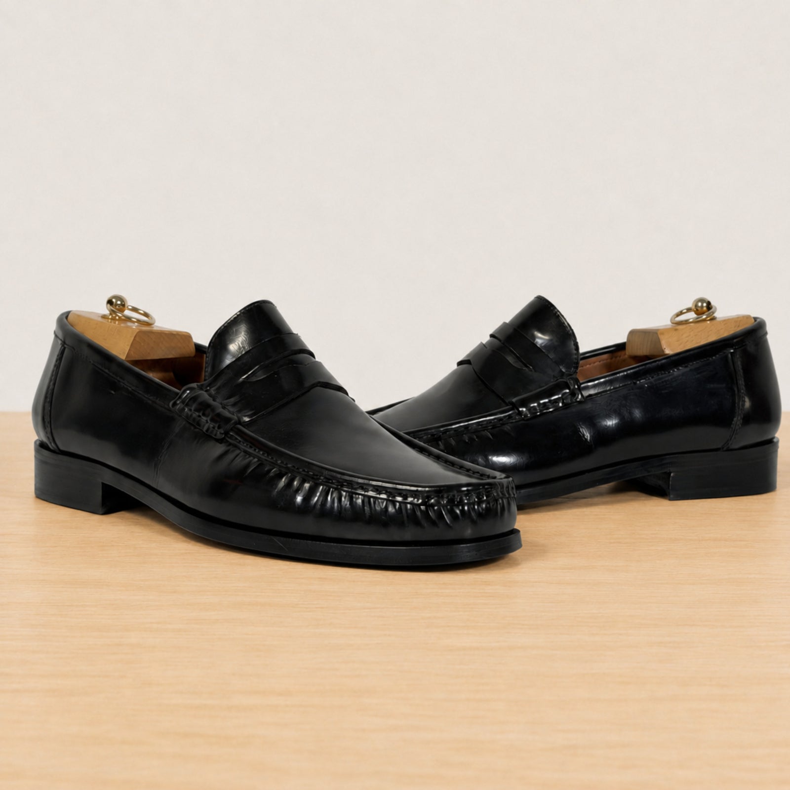 A pair of glossy black leather penny loafers with decorative stitching across the vamp, displayed on a light wood surface against a plain off-white background.