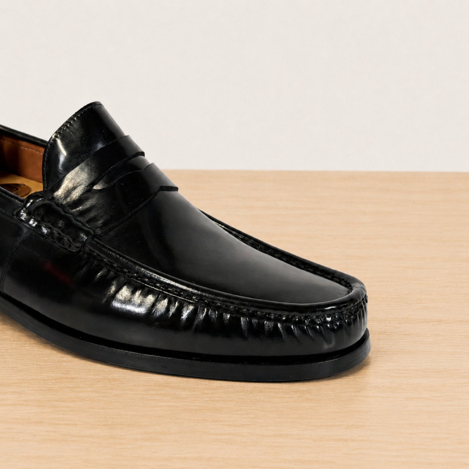 A close-up of a glossy black leather penny loafer on a light wood surface against a neutral off-white background.