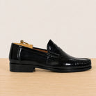 A single black leather penny loafer shown in profile on a light wood surface against a plain off-white background.