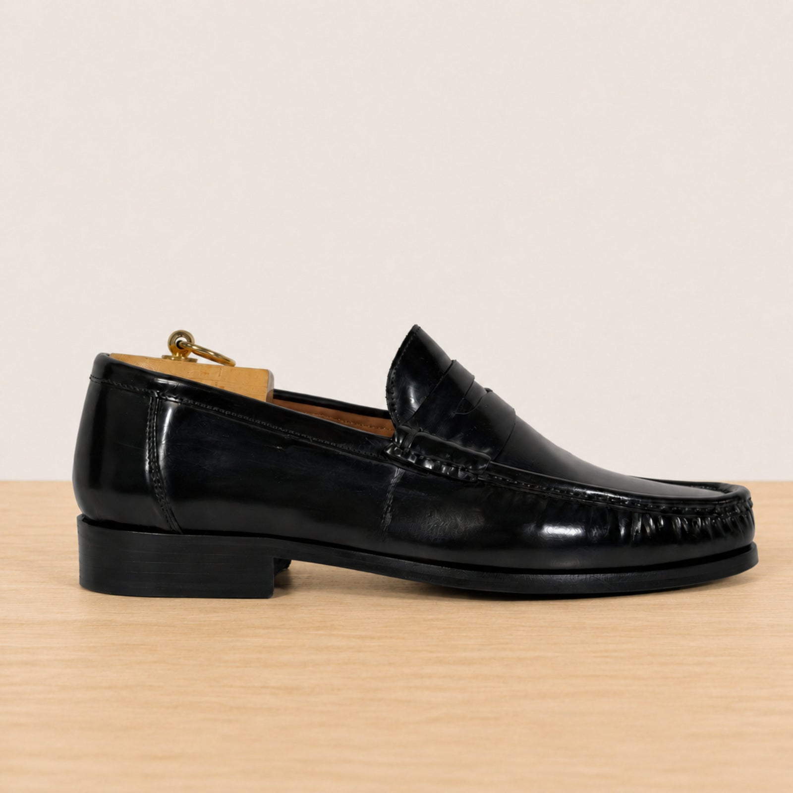 A single black leather penny loafer shown in profile on a light wood surface against a plain off-white background.