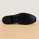 The outsole of black leather dress shoe sole, showing clean stitching and a small embossed brand mark, resting on a light wood surface against a neutral off-white background.