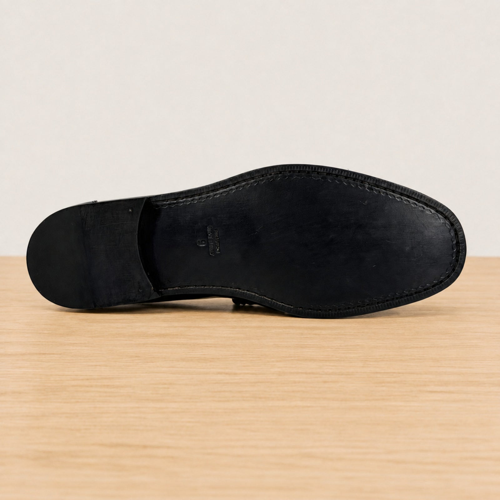 The outsole of black leather dress shoe sole, showing clean stitching and a small embossed brand mark, resting on a light wood surface against a neutral off-white background.