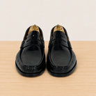 A pair of black leather penny loafers with decorative stitching, displayed on wooden shoe trees on a light wood surface against a cream background.