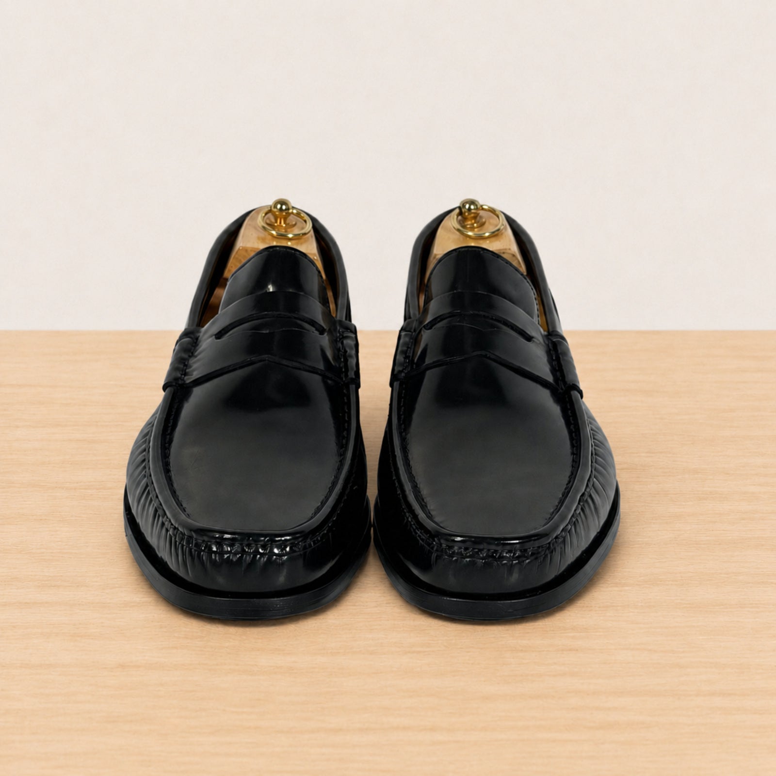 A pair of black leather penny loafers with decorative stitching, displayed on wooden shoe trees on a light wood surface against a cream background.