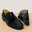 A pair of black leather penny loafers with decorative stitching and low block heels, displayed on a light wood surface against a neutral off-white background. One shoe is shown from a front three-quarter angle on a wooden shoe tree, and the other is shown from a side profile angle.