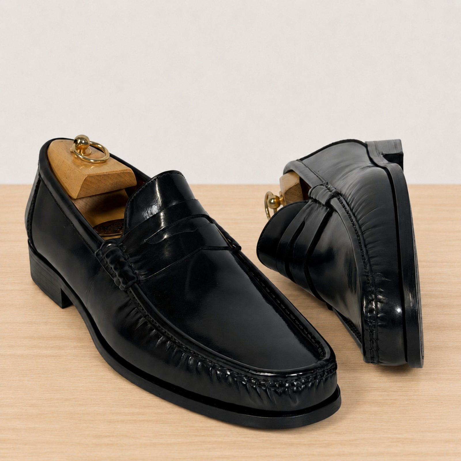 A pair of black leather penny loafers with decorative stitching and low block heels, displayed on a light wood surface against a neutral off-white background. One shoe is shown from a front three-quarter angle on a wooden shoe tree, and the other is shown from a side profile angle.