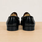A pair of black leather loafers shown from the back, featuring a polished finish, set on a light wood surface against a plain off-white background.