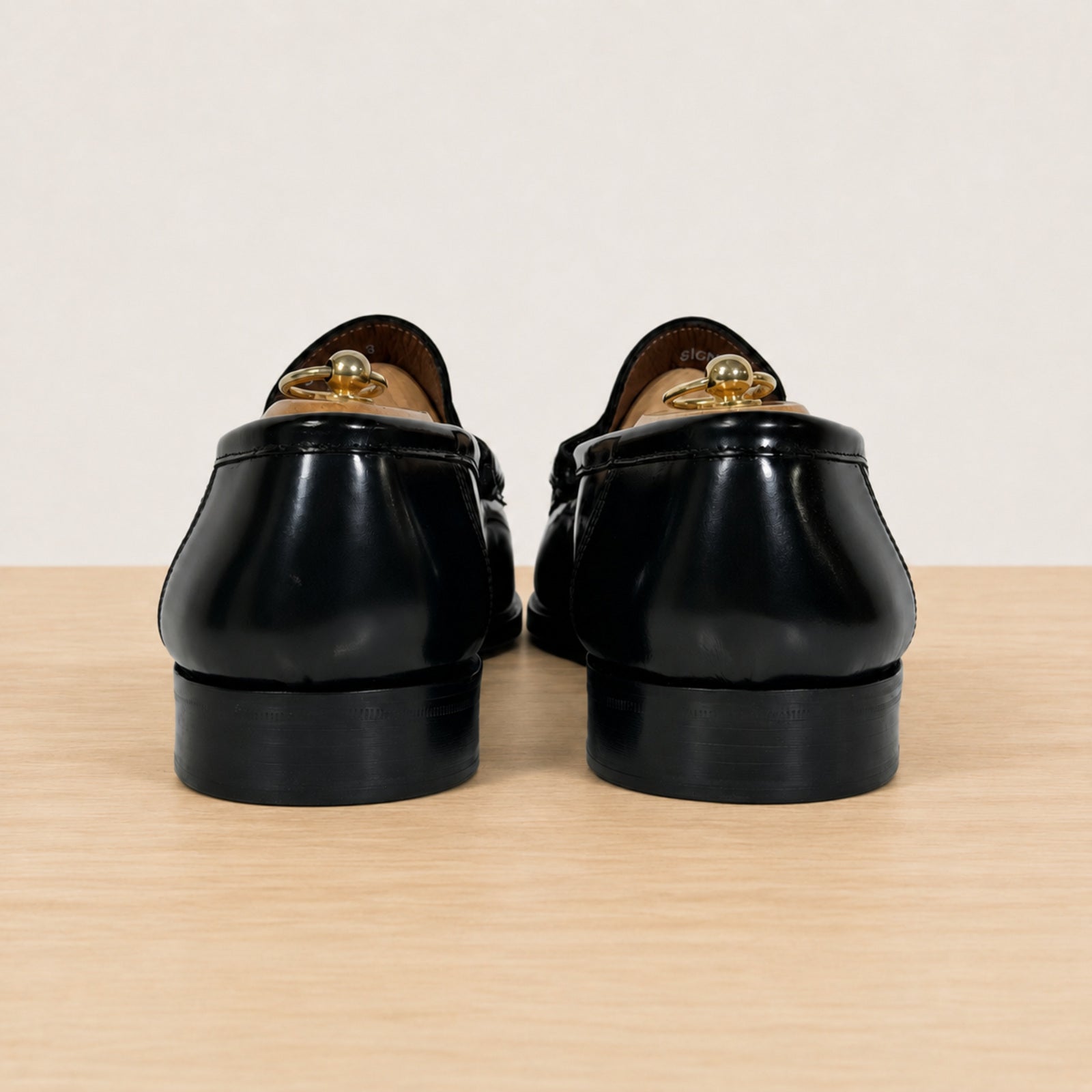 A pair of black leather loafers shown from the back, featuring a polished finish, set on a light wood surface against a plain off-white background.