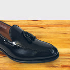 A close-up of a Black leather tassel loafer with a leather sole, shown from a front angle on a wooden table.