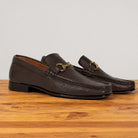A pair of dark brown leather loafer with a gold buckle on a wooden surface