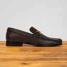 A dark brown  leather loafer with a gold-tone horsebit detail on the vamp, displayed in profile on a wooden surface against a light gray background.