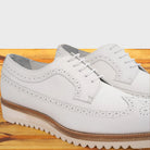 A pair of white leather wingtip oxford shoes with decorative brogue perforations, resting on a light-colored wooden surface.