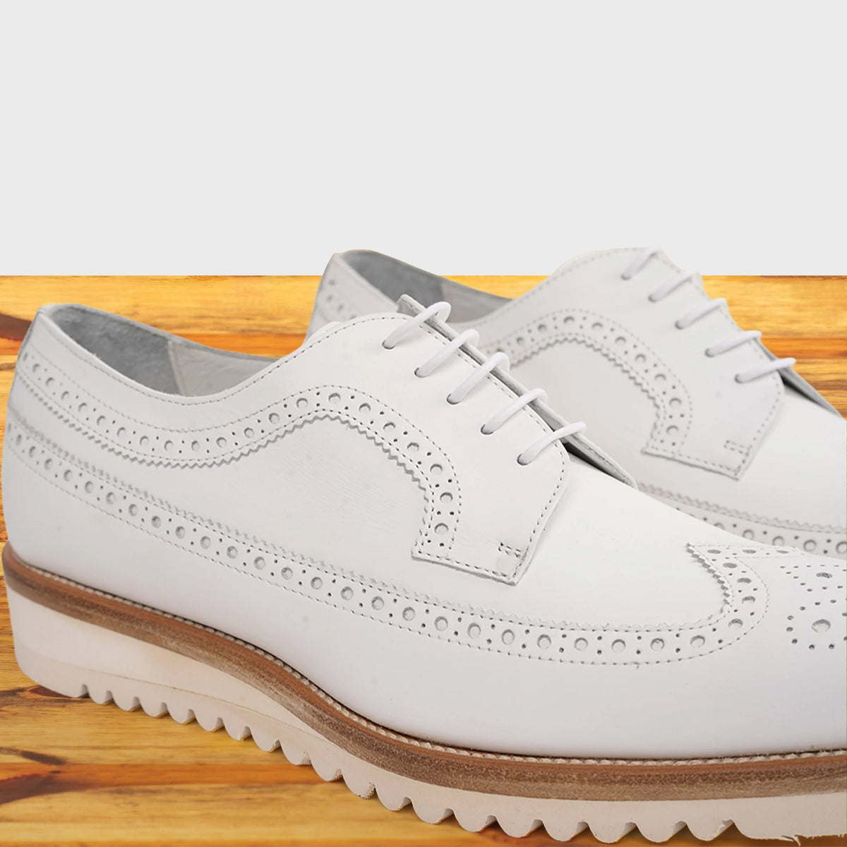A pair of white leather wingtip oxford shoes with decorative brogue perforations, resting on a light-colored wooden surface.