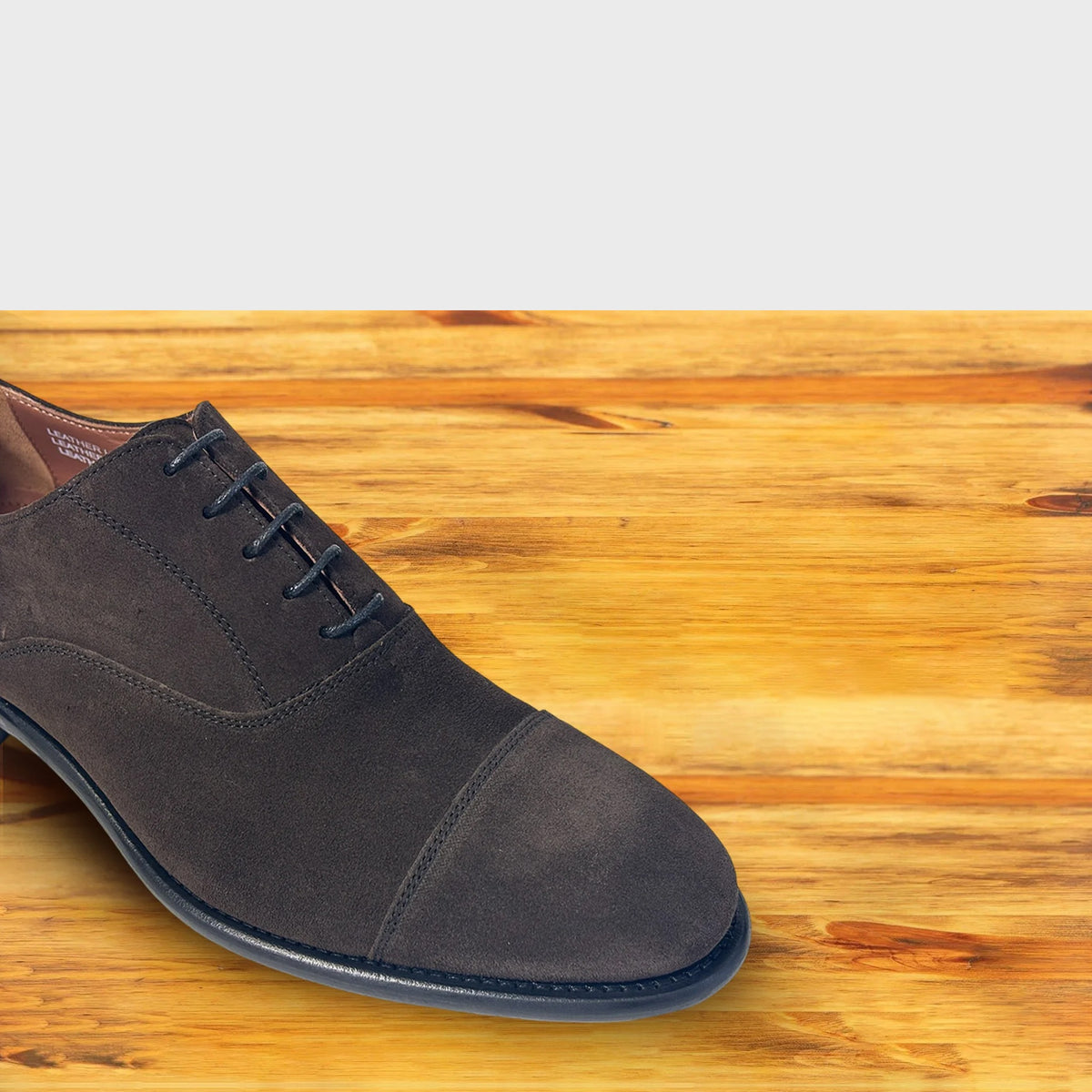 A Dusty Brown suede Oxford cap-toe shoe with a leather sole, displayed on a wooden surface.