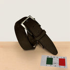 A dark brown suede belt with a silver buckle, coiled on a light wood surface against a white background, with a 'MADE IN ITALY' card in the foreground.