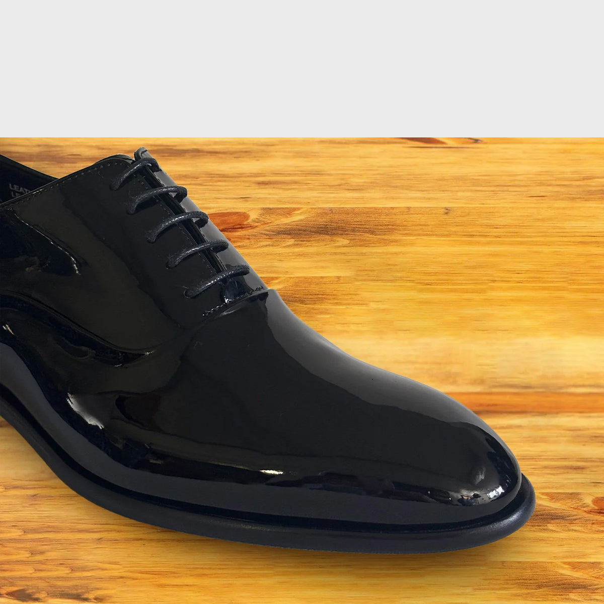 A glossy black patent leather oxford dress shoe with black laces, shown from a high angle on a light brown wooden.