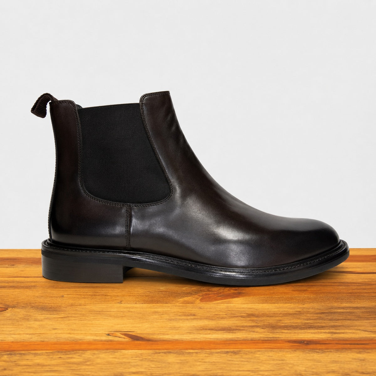 Dark Brown leather Chelsea boot on a wooden surface with a light gray background