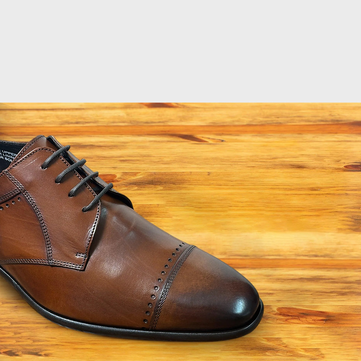 Mahogany leather cap-toe shoe, close-up of the toe and lacing showing the burnished finish, and black leather sole on a wooden surface