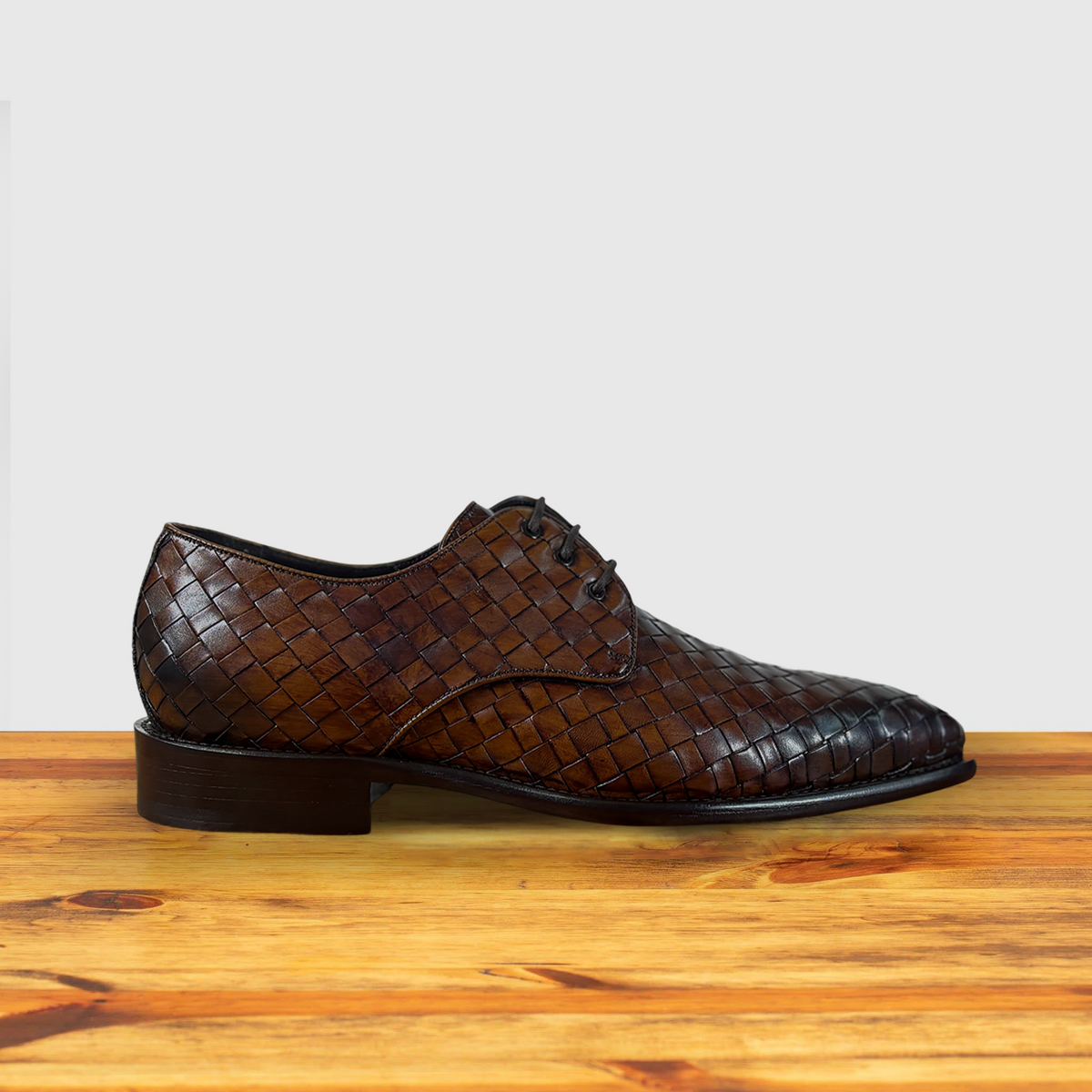 Mahogany woven leather shoe on a wooden surface with a light gray background