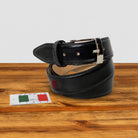A Graphite leather belt with a silver buckle, coiled on a wooden surface next to a small 'Made in Italy' card.