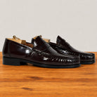 A pair of glossy burgundy penny loafers with a high-shine patent finish, displayed on a wooden surface against a neutral light gray background.