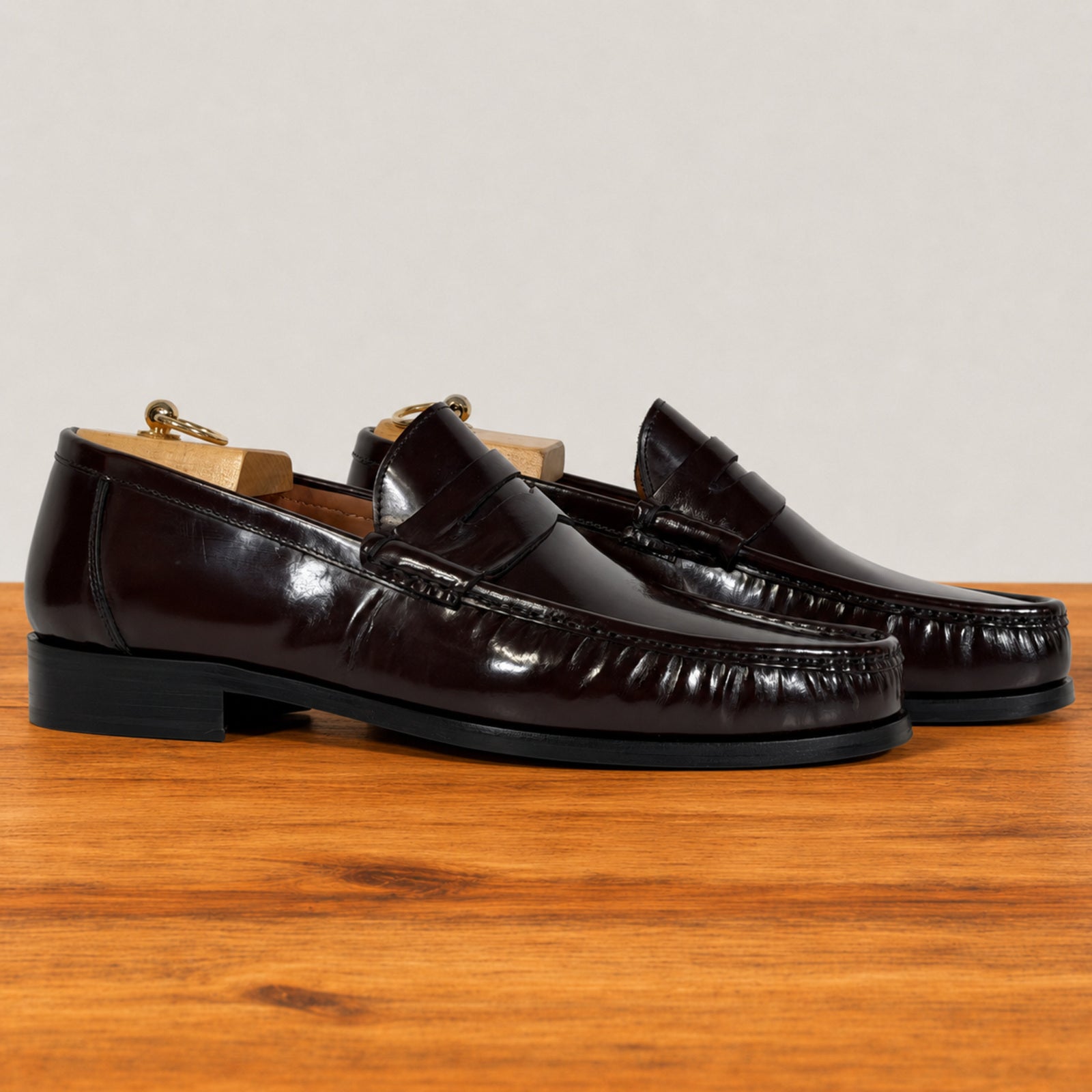 A pair of glossy burgundy penny loafers with a high-shine patent finish, displayed on a wooden surface against a neutral light gray background.