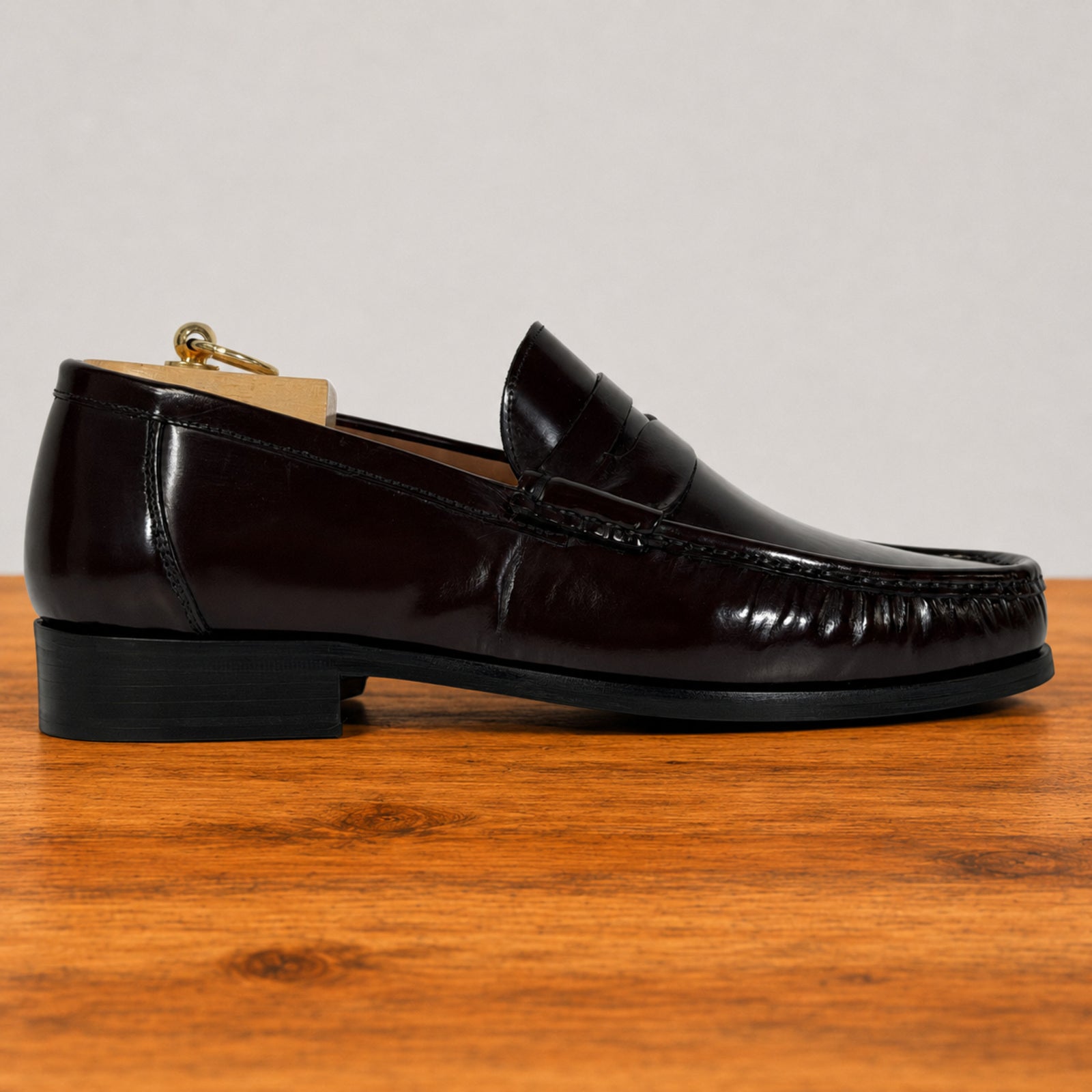 A single burgundy glossy leather penny loafer with a black sole, shown in profile on a wooden surface against a light gray background.