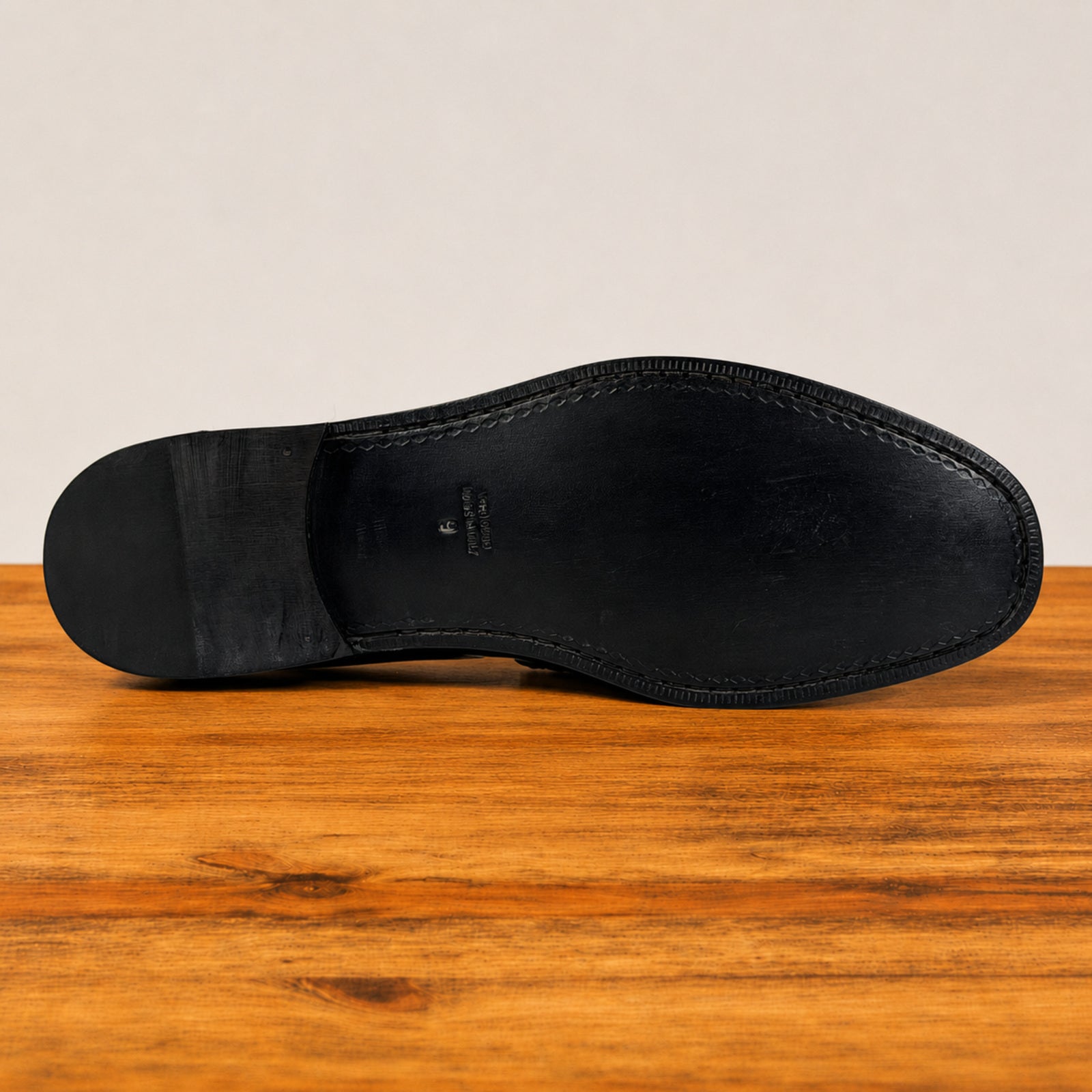 The black leather sole of a dress shoe, showing the heel, vamp, and toe, resting on a wooden surface against a light beige background.