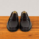 A pair of glossy burgundy leather penny loafers with decorative stitching, displayed on wooden shoe trees on a wooden surface against a light gray background.