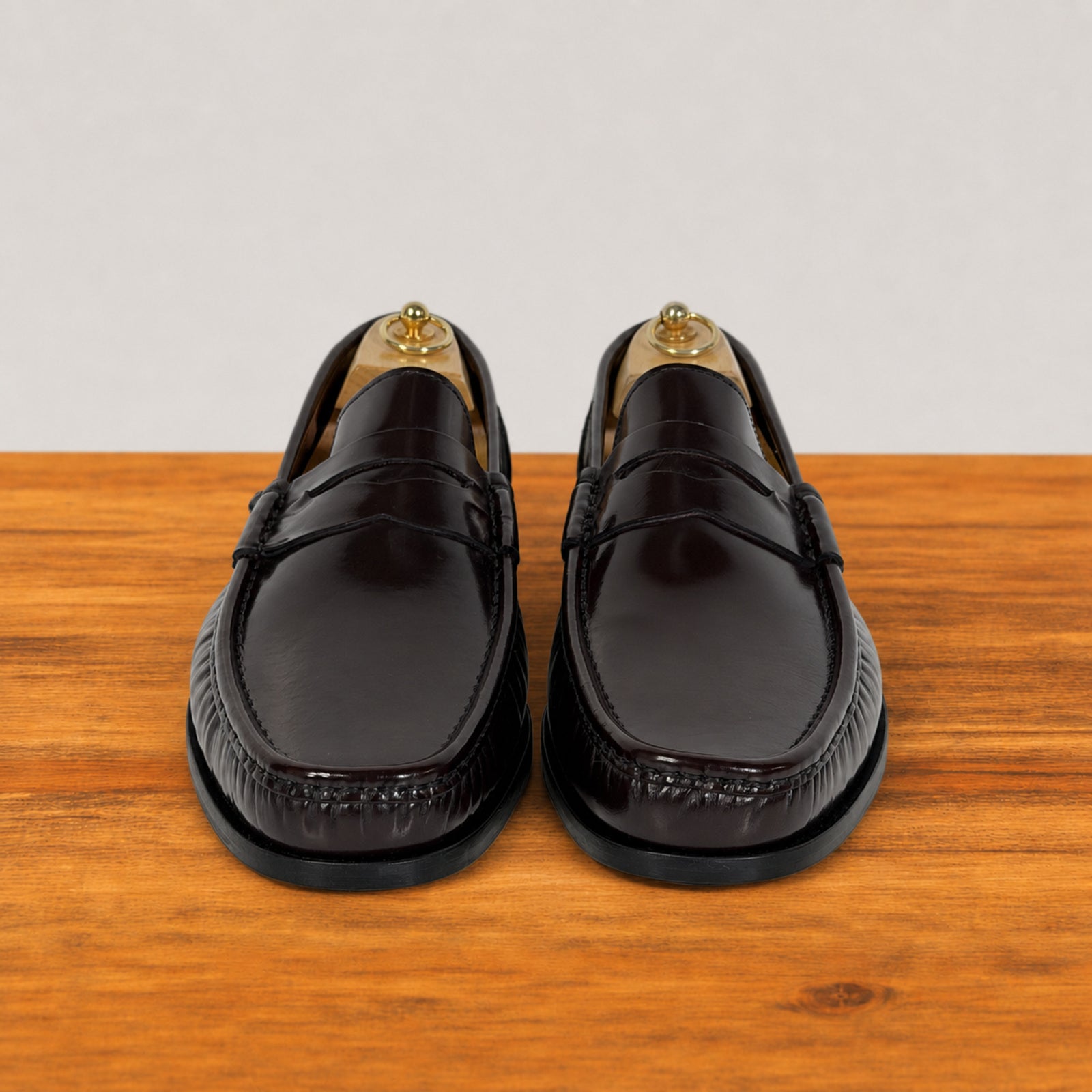 A pair of glossy burgundy leather penny loafers with decorative stitching, displayed on wooden shoe trees on a wooden surface against a light gray background.