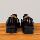 A pair of burgundy patent leather penny loafers, shown from the back on a wooden surface against a light gray background.