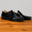 A pair of glossy black leather penny loafers displayed on wooden shoe trees over a polished wooden surface against a light gray background.