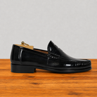 A single black leather penny loafer shoe with a low stacked heel, shown in profile on a wooden surface against a light gray background.