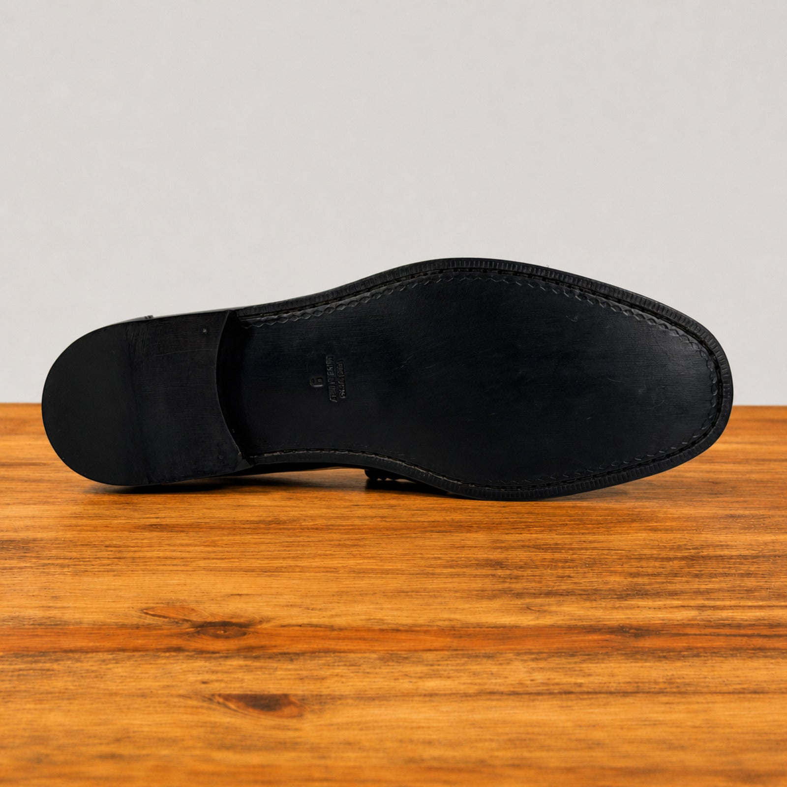 The outsole of a black leather shoe is shown from a top-down view, resting on a wooden surface against a light gray background.