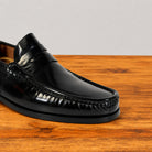 A close-up of a black leather penny loafer shoe resting on a wooden surface against a light gray background.