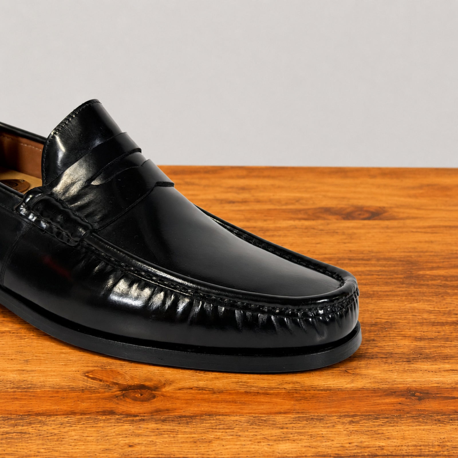 A close-up of a black leather penny loafer shoe resting on a wooden surface against a light gray background.