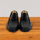 A pair of black leather penny loafers with decorative stitching and tassel details, displayed on wooden shoe trees on a wooden surface against a light gray background.