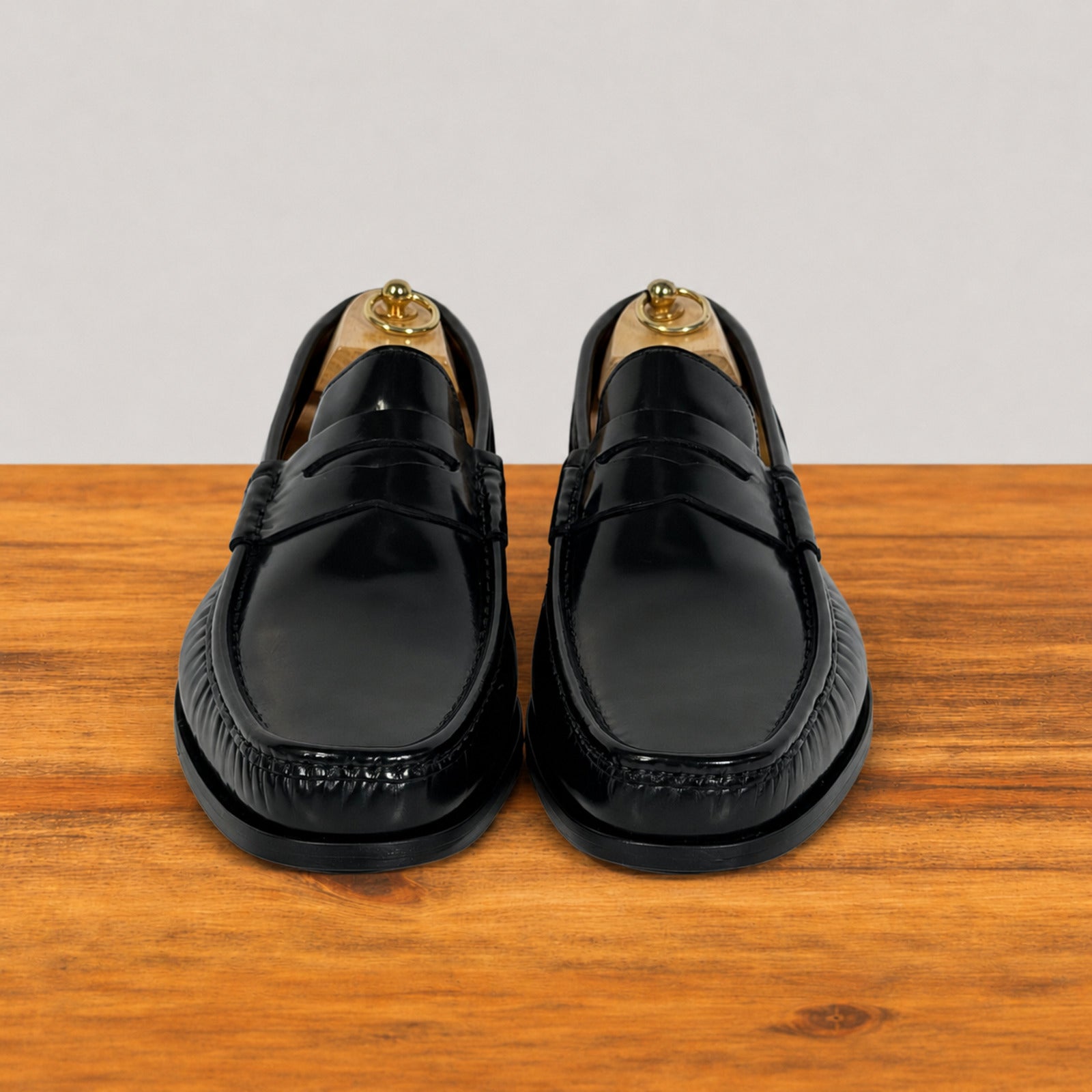 A pair of black leather penny loafers with decorative stitching and tassel details, displayed on wooden shoe trees on a wooden surface against a light gray background.