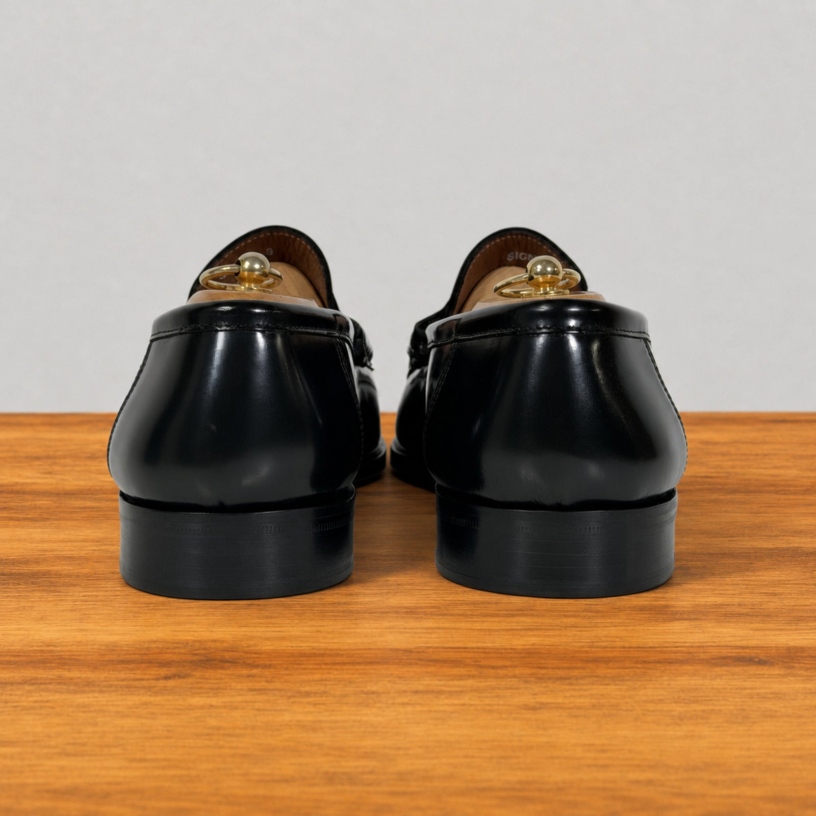 A pair of black leather loafers shown from the back, resting on a wooden surface against a light gray background. 