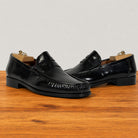 A pair of black leather penny loafers with decorative stitching and low block heels, displayed on wooden wooden surface against a light gray background.