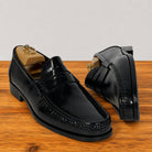 A pair of black leather penny loafers displayed on a wooden surface against a light gray background. One shoe is shown from the front, resting on a wooden shoe tree, and the other is positioned behind it showing the side and heel.