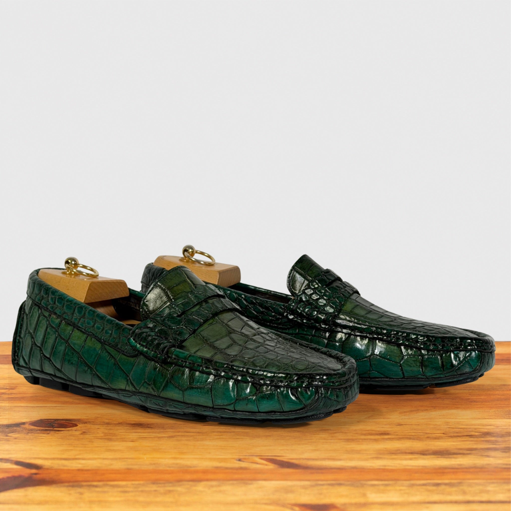 A pair of Emerald Green crocodile-embossed leather penny loafers resting on a wooden surface with wooden shoe trees inside, against a plain light gray background.