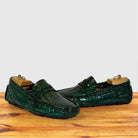 A pair of Emerald Green crocodile-embossed leather loafers, displayed on wooden shoe trees on a light wood surface against a plain light gray background.