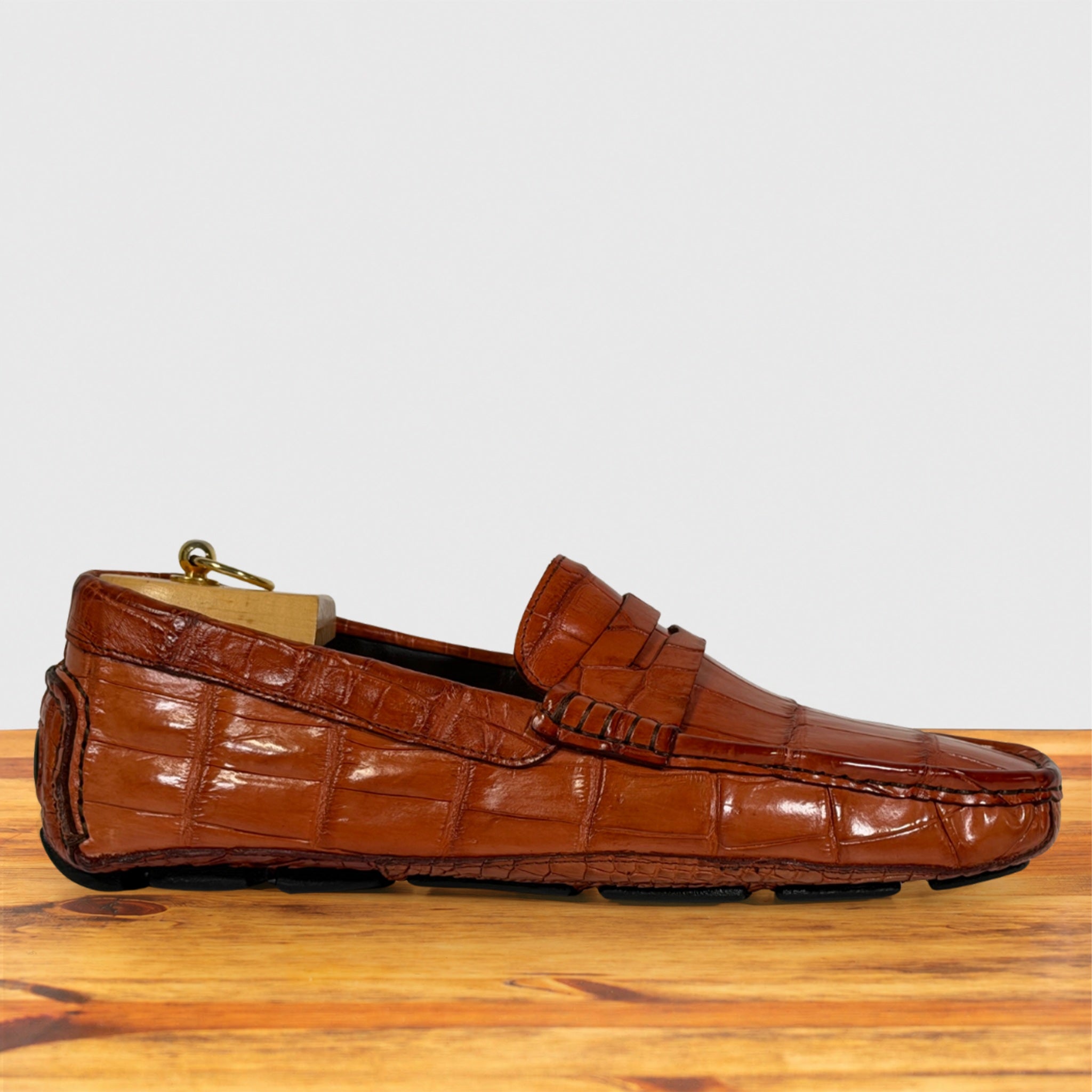 A single Burnt Orange crocodile-embossed leather loafer driving shoe, resting on a wooden surface against a light gray background.