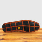 The outsole of a brown crocodile-embossed leather shoe resting on a wooden surface against a light gray background.