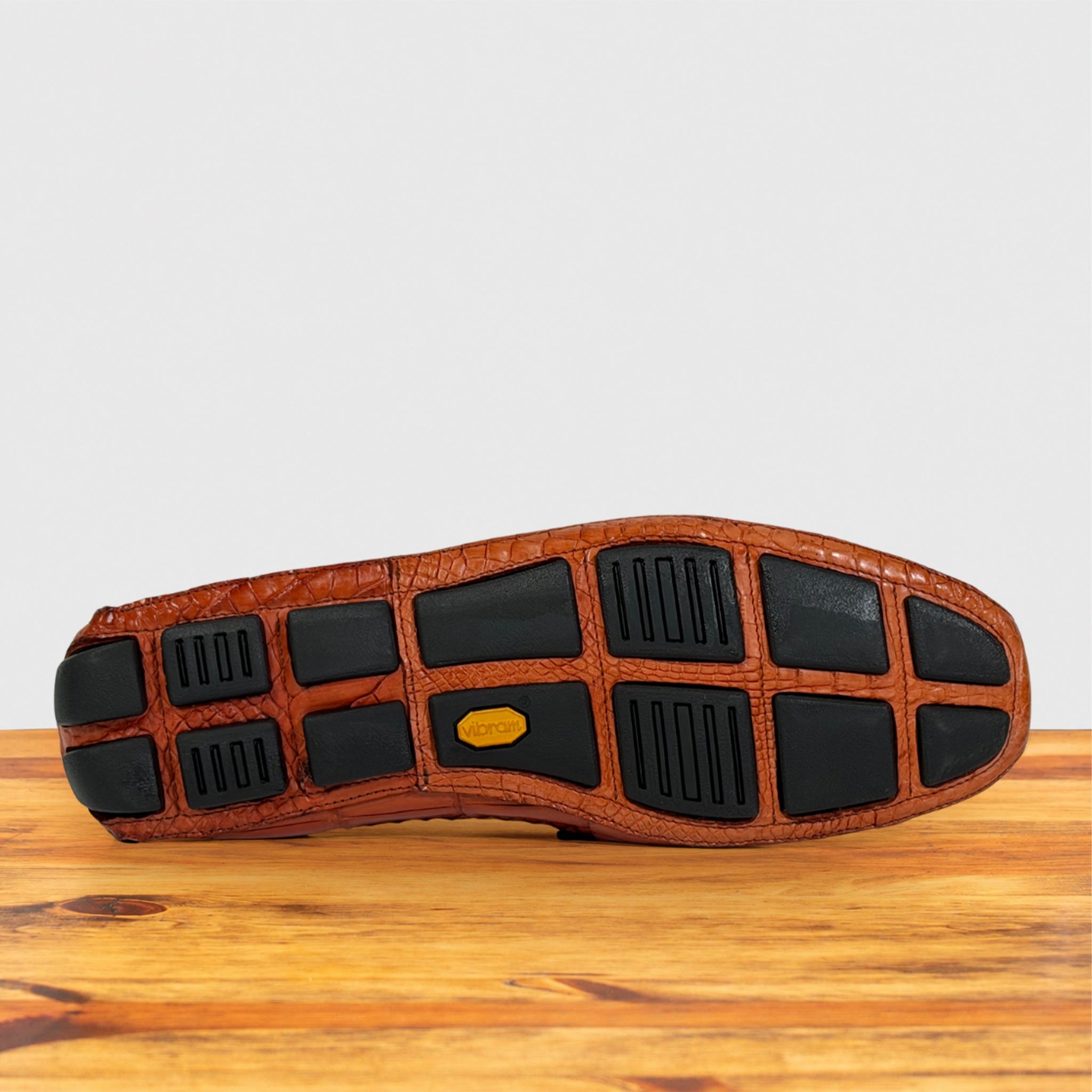 The outsole of a brown crocodile-embossed leather shoe resting on a wooden surface against a light gray background.
