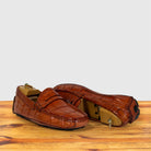 A pair of Burnt Orange crocodile-embossed leather penny loafers displayed on a wooden surface against a light gray background.