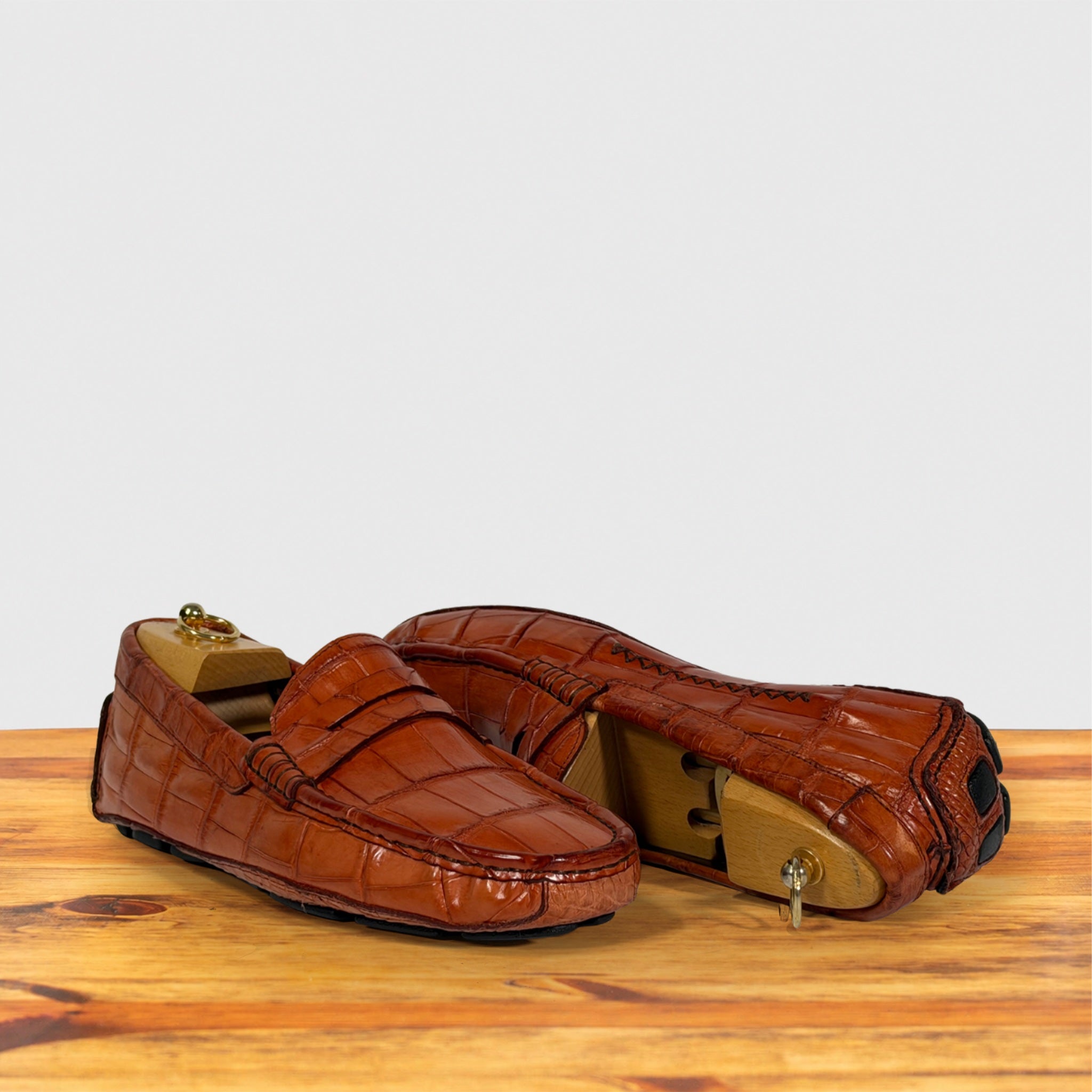 A pair of Burnt Orange crocodile-embossed leather penny loafers displayed on a wooden surface against a light gray background.